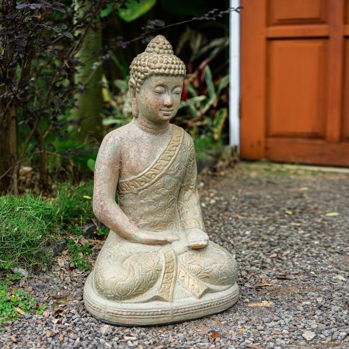 A seated Buddha garden statue in a light beige stone or concrete finish, placed outdoors on a gravel path with green plants and a reddish-brown wooden door in the background.