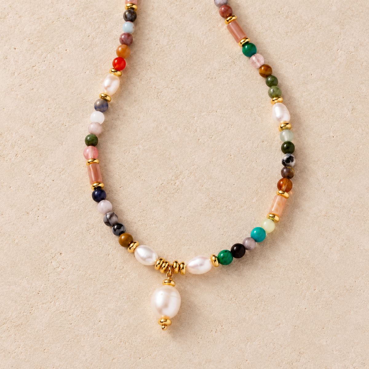 A beaded necklace with white baroque pearls, colorful stone beads, and gold spacer rings, featuring a large white pearl pendant hanging from a gold clasp, displayed on a textured beige background.