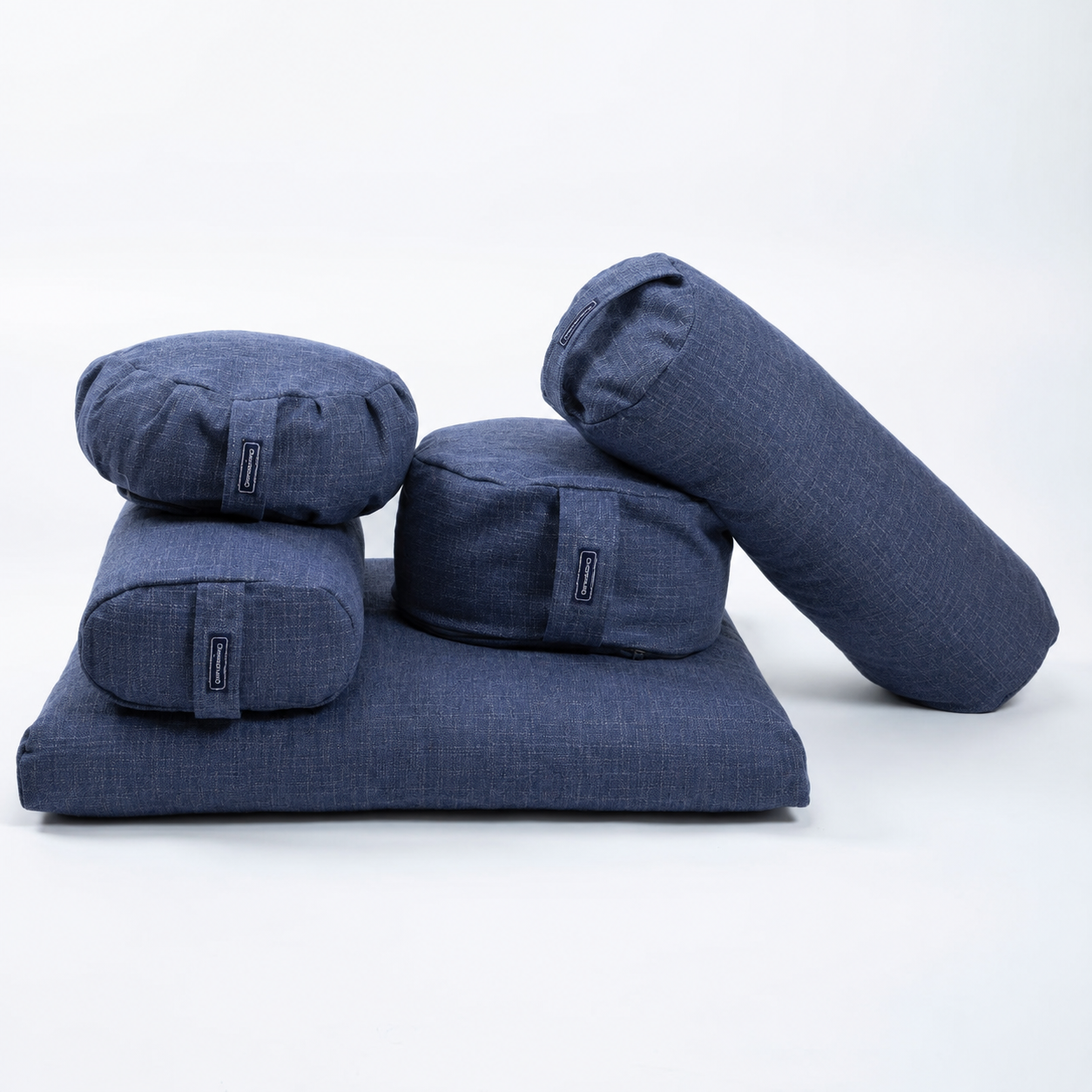 A set of five navy blue fabric yoga props, including a rectangular floor cushion, three cylindrical bolsters, and two round support pillows, arranged on a seamless white background.