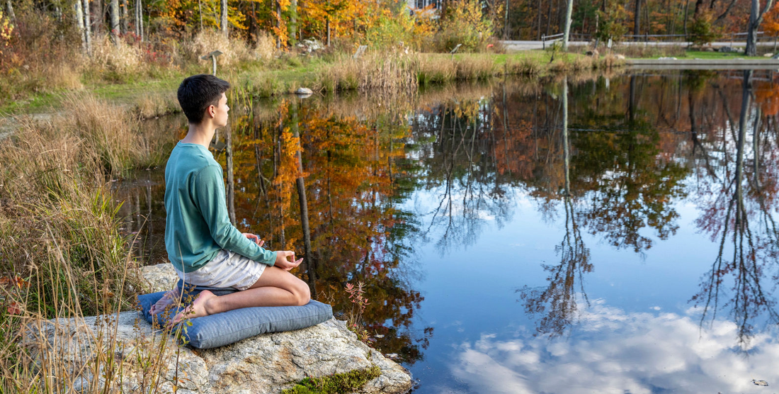 boy sitting on zabuton meditation cushion on top of a rock over looking a pond with fall leaves and trees outside