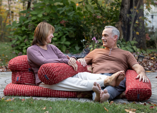 man and woman sitting outside with fall leaves resting on meditation bolsters and zabu and zafuton set with trees in the background