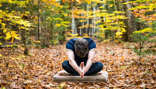 man sitting on zafu & zabuton meditation cushion outside in autumn leaves meditating while bowing