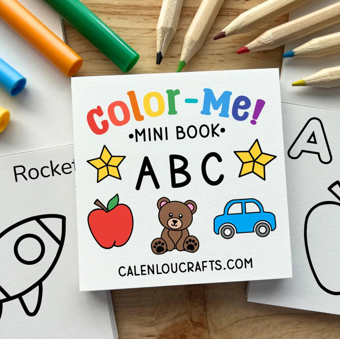A children's mini coloring book titled 'Color-Me! Mini Book ABC' shown on a wooden surface with colored pencils and other coloring pages around it. The cover features colorful title text, yellow stars, an apple, a teddy bear, and a blue car.