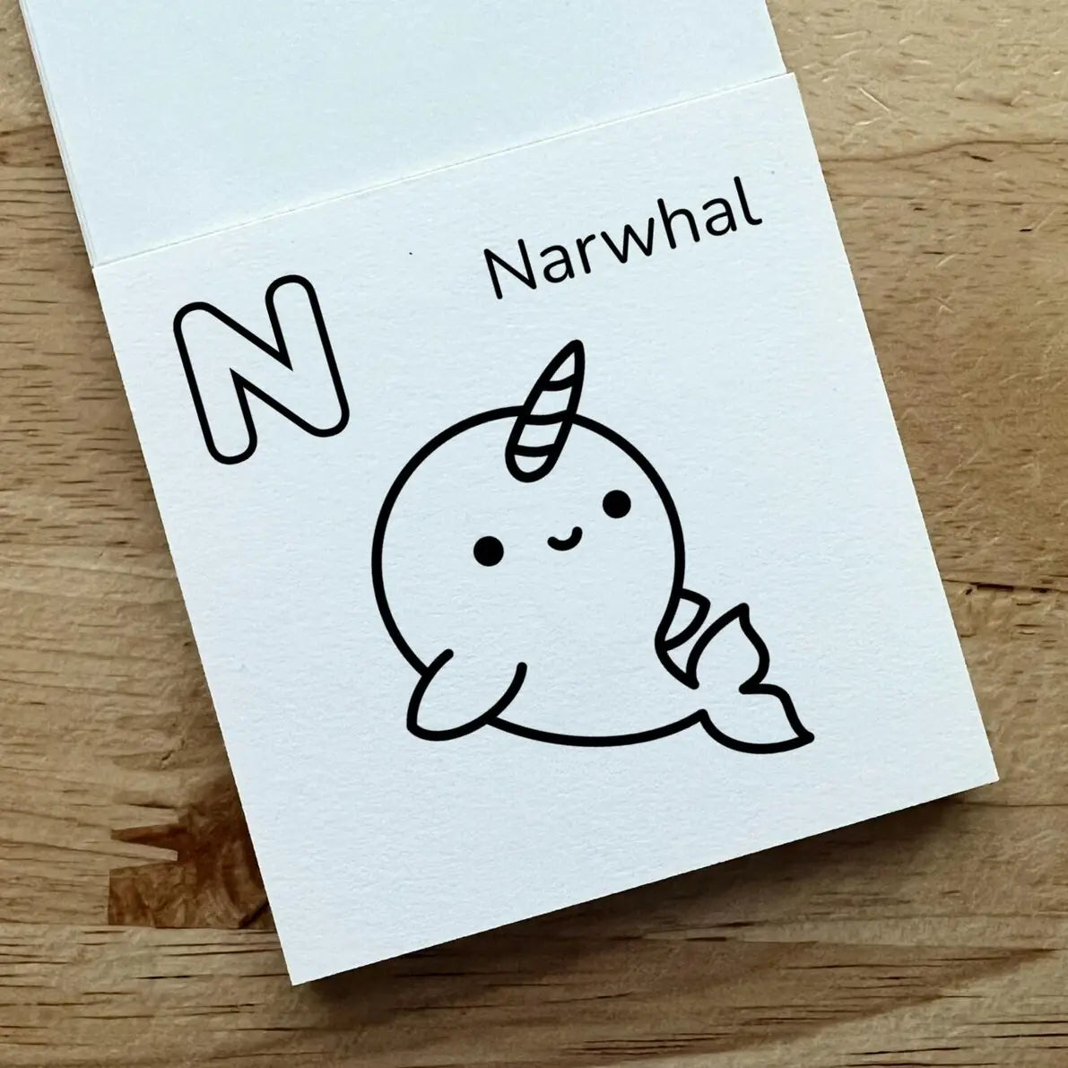 A white alphabet card featuring the letter 'N' and the name 'Narwhal' above a simple black line drawing of a narwhal, photographed on a light wood surface.