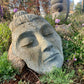 Stone sculpture of a face in a garden setting with plants and flowers.