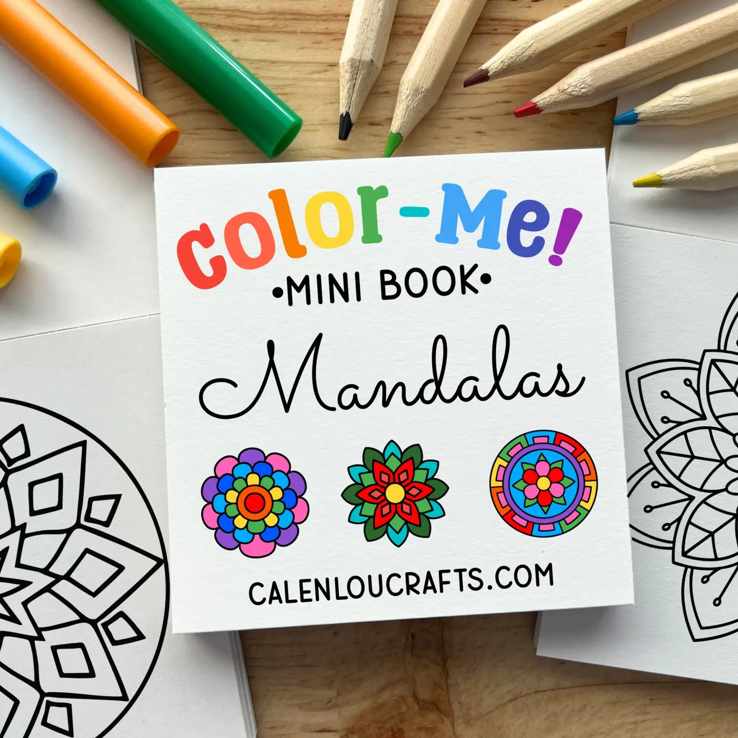 A square white booklet titled "color-Me! Mini Book: Mandalas" lies on a wooden surface, surrounded by colored pencils, markers, and partially filled coloring pages with mandala designs.