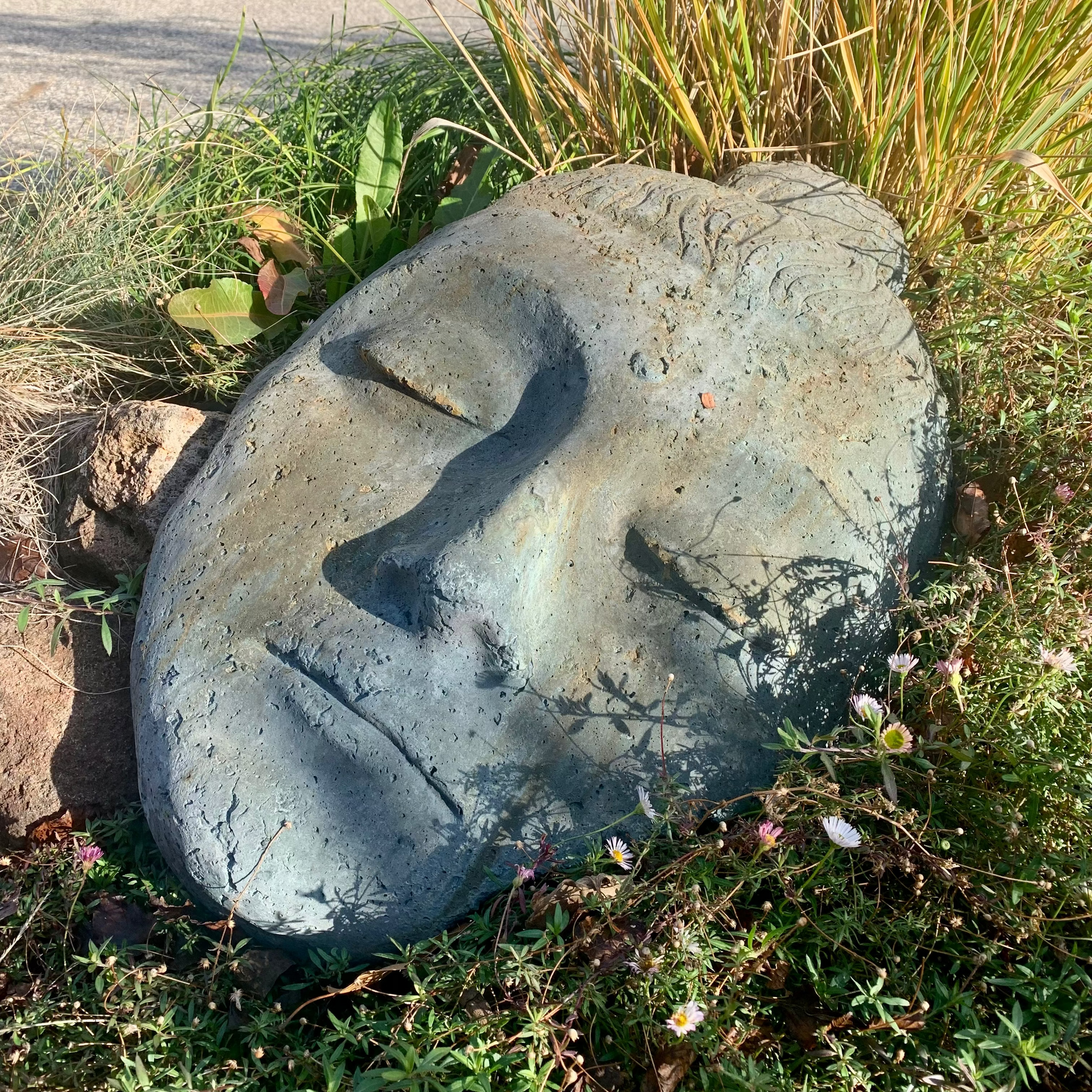 Concrete sculpture of a face in a natural setting with grass and plants.