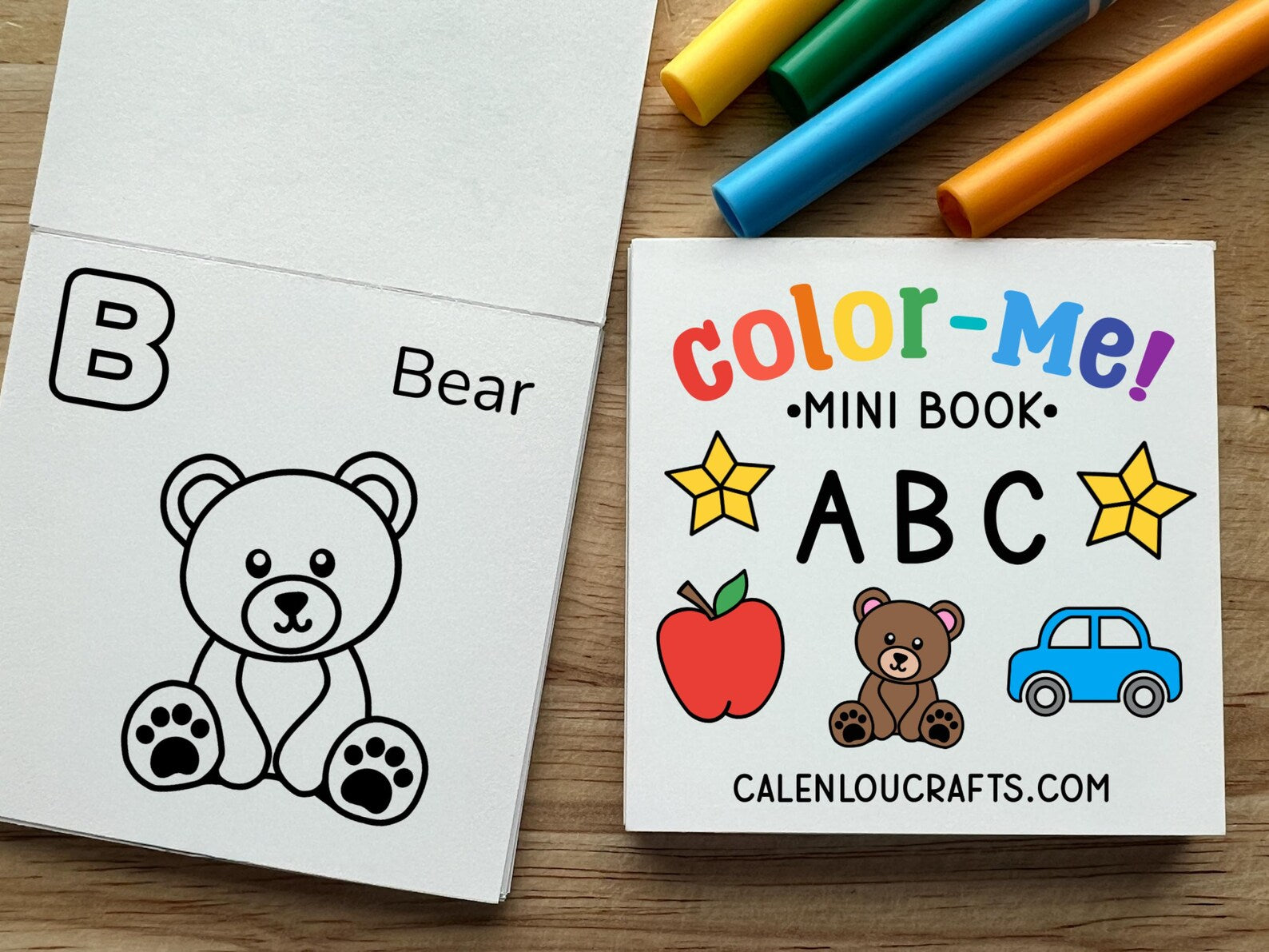 A children's mini ABC coloring book shown open on a wooden surface, alongside the front cover and colorful markers. The book features black-and-white bear illustrations on the inside pages and a colorful cover with the title 'Color-Me! Mini Book ABC' and the brand name CALENOUCRAFTS.