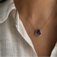 A close-up of a person wearing a delicate gold chain necklace with a teardrop-shaped purple gemstone pendant, styled with a white linen shirt.