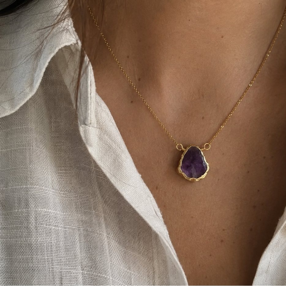 A close-up of a person wearing a delicate gold chain necklace with a teardrop-shaped purple gemstone pendant, styled with a white linen shirt.