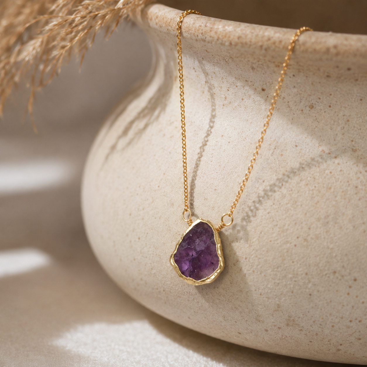 A gold chain necklace with a raw purple amethyst geode pendant, resting on the rim of a textured beige ceramic bowl with dried grass in the background.