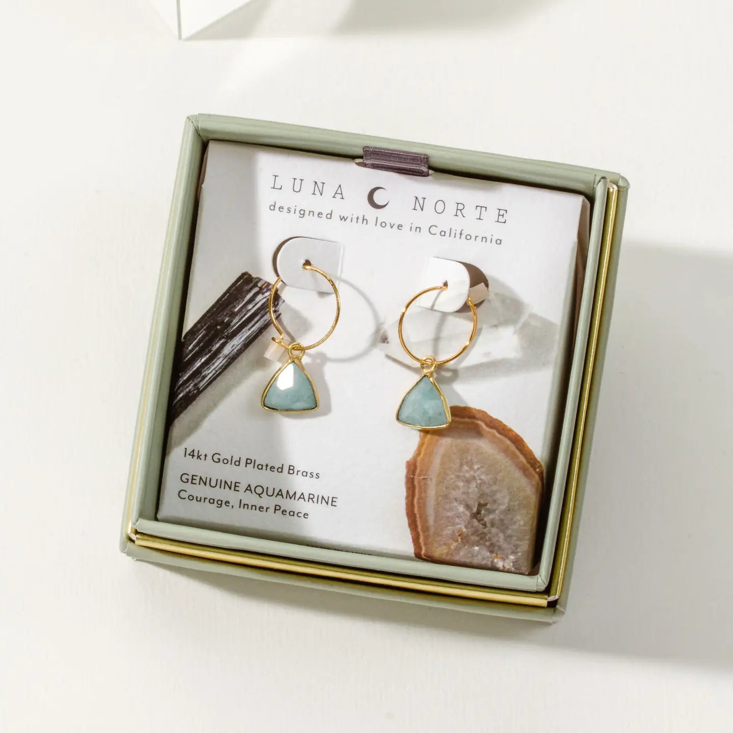 A pair of gold-plated brass hoop earrings with triangular aquamarine stones, presented in a square gift box by Luna Norte. The earrings are displayed on a white backing card with mineral samples as props, set against a clean white background.