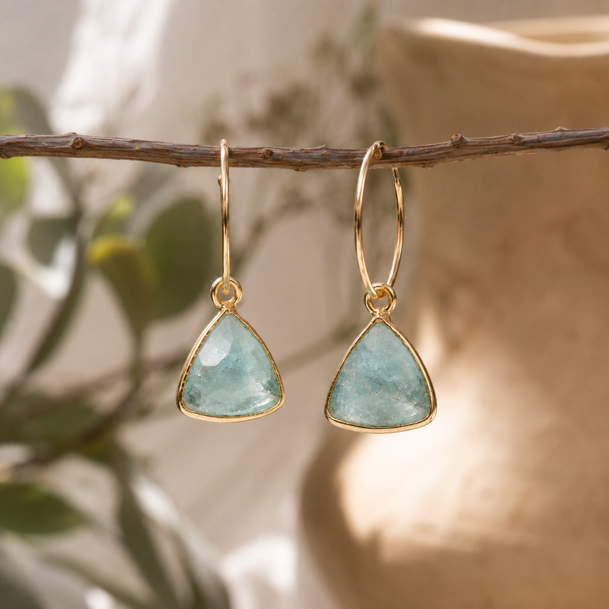 A pair of gold hoop earrings with triangular aquamarine blue-green stone drops, hanging from a thin brown branch against a softly blurred beige ceramic vase and green foliage background.