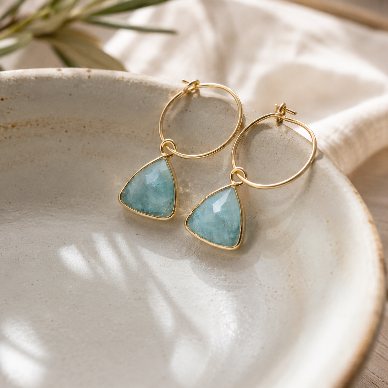Gold hoop earrings with triangular light blue stone pendants, displayed on a textured ceramic dish with soft neutral fabric in the background.