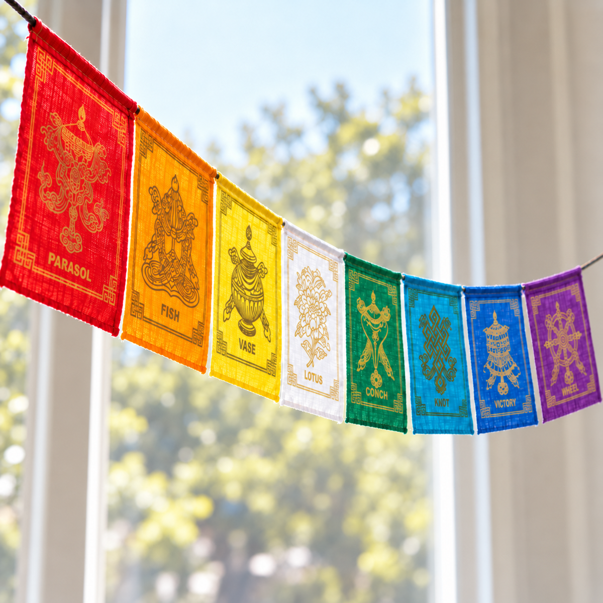 A string of colorful Tibetan prayer flags hangs in front of a bright window with blurred green trees outside. The flags are red, orange, yellow, white, green, blue, and purple, each with gold traditional symbols and Sanskrit mantras.