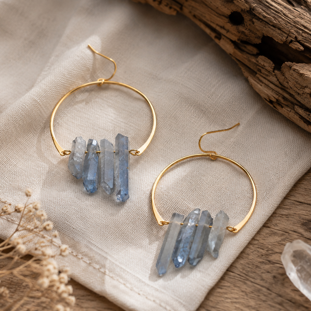 A pair of gold-tone hoop earrings with dangling natural blue crystal points, displayed on a beige linen cloth with dried flowers and rustic wood in the background.