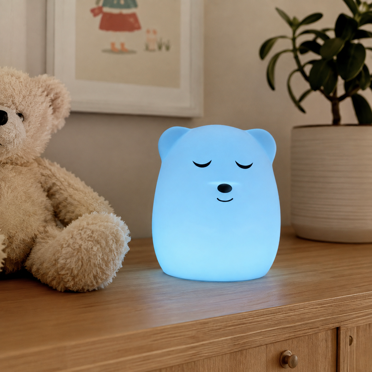 A cute bear-shaped night light glowing with a soft blue light, placed on a wooden dresser next to a teddy bear and a potted plant.