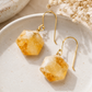 A pair of gold-tone drop earrings with faceted yellow-orange crystal pendants, displayed on a textured beige ceramic plate with dried flowers in the background.