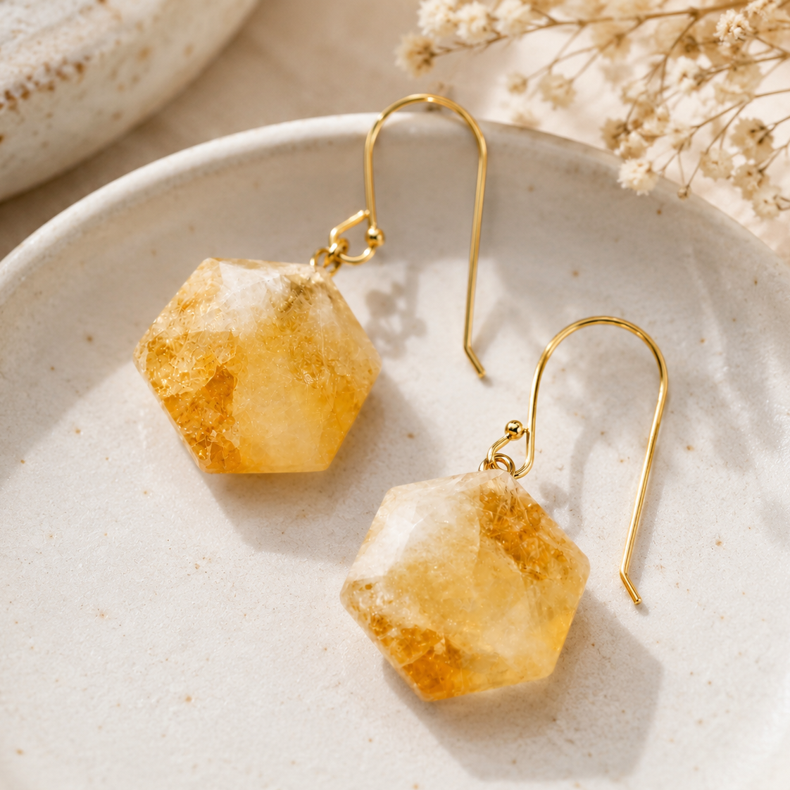 A pair of gold-tone drop earrings with faceted yellow-orange crystal pendants, displayed on a textured beige ceramic plate with dried flowers in the background.
