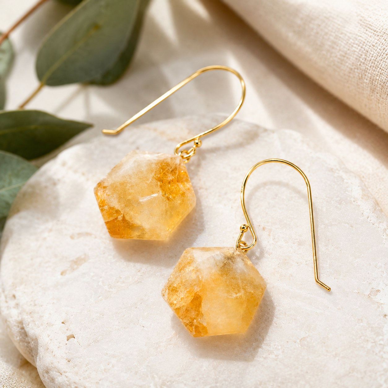 A pair of gold-tone dangle earrings with faceted, translucent amber-orange gemstone pendants, displayed on a textured white stone slab with green leaves and a beige fabric background.