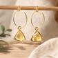A pair of gold hoop dangle earrings with faceted yellow triangular gemstones, hanging from a wooden rod against a softly blurred neutral background.