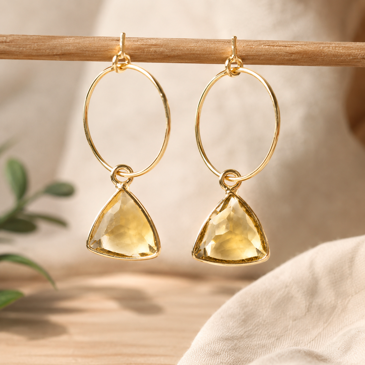 A pair of gold hoop dangle earrings with faceted yellow triangular gemstones, hanging from a wooden rod against a softly blurred neutral background.
