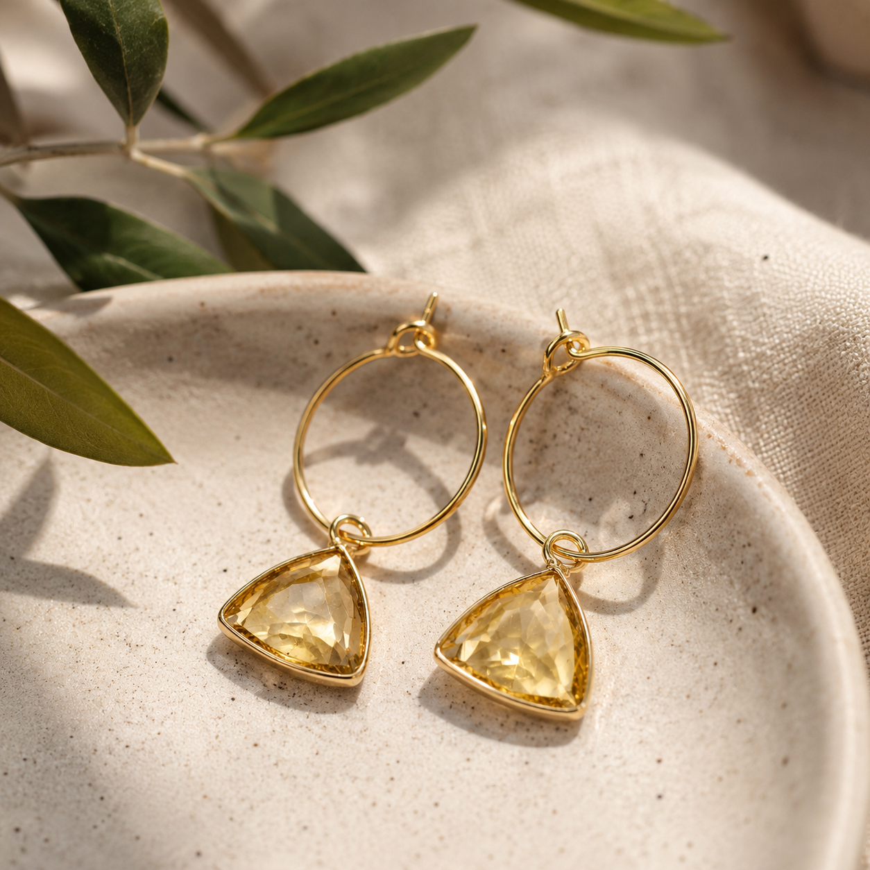 A pair of gold hoop earrings with triangular yellow gemstone pendants, resting on a textured beige ceramic dish with green leaves in the background.
