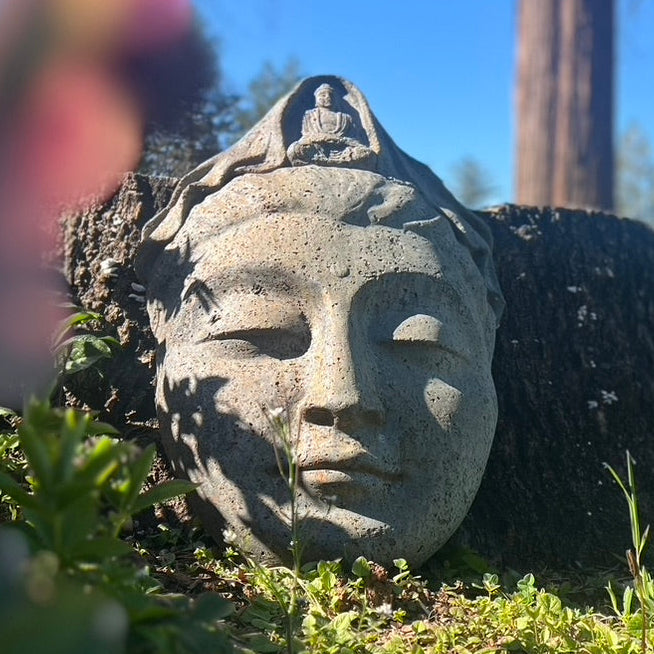 Stone sculpture of Kwan Yin with a smaller figure on top, set against a natural background.