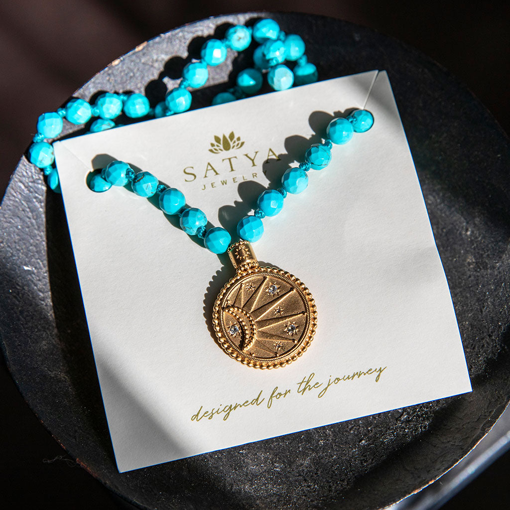 Turquoise beads and gold pendant mala resting on a black wood bowl.