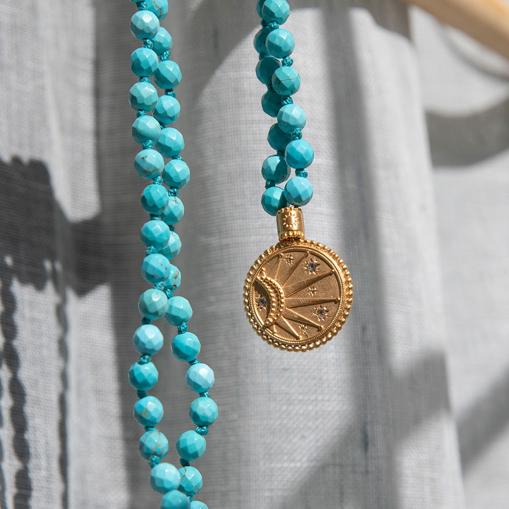 Turquoise beads and gold pendant mala hanging on a wood beam.