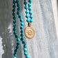 Turquoise beads and gold pendant mala resting up against a white cloth.