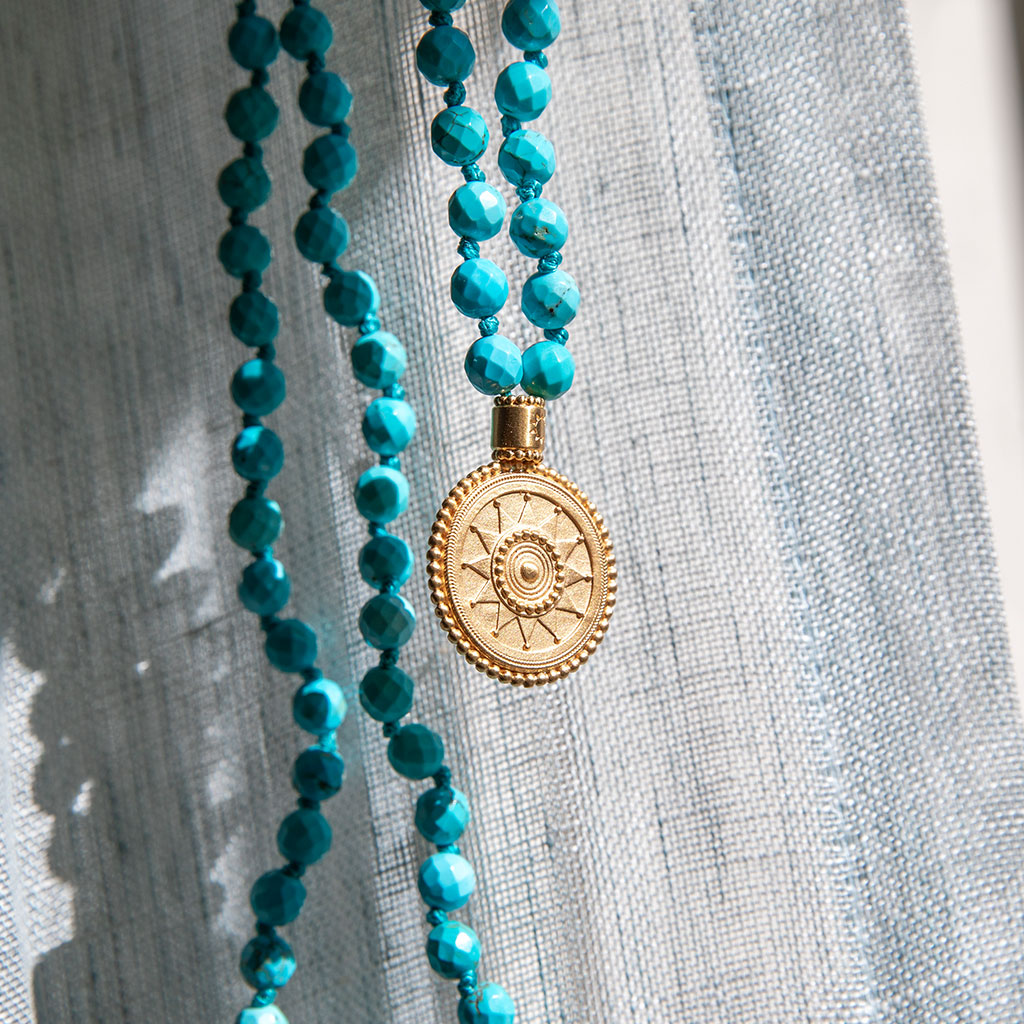 Turquoise beads and gold pendant mala resting up against a white cloth.