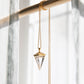 Crystal pendant on a gold necklace hanging on a wod beam up against a white curtain.