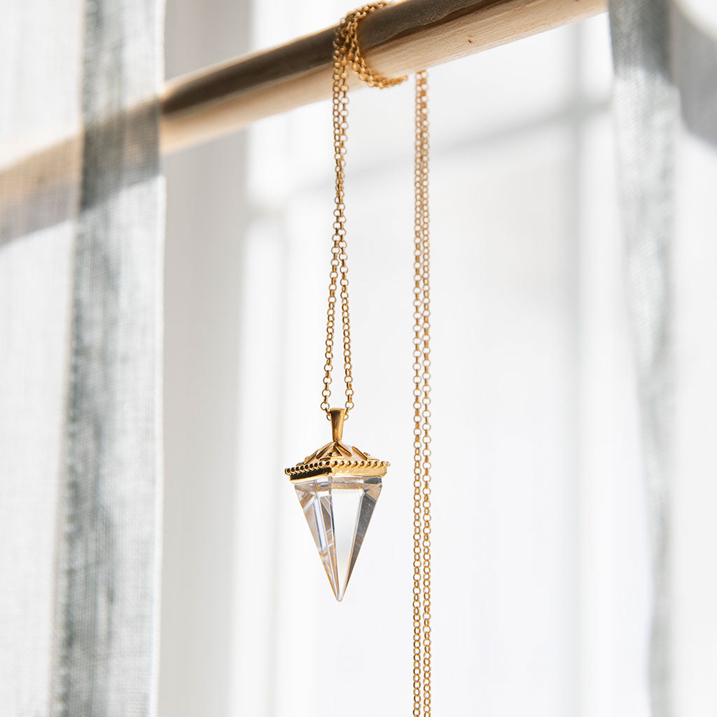 Crystal pendant on a gold necklace hanging on a wod beam up against a white curtain.