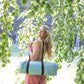 A woman standing outdoors among leafy branches, carrying a rolled light blue yoga mat over her shoulder.