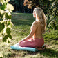 A woman with long blonde hair sits cross-legged on a light blue cushion on a grassy lawn, viewed from behind and side, wearing a beige top and pink pants, framed by green leaves in the foreground.