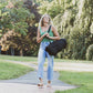 A woman walking barefoot on a paved path in a park, carrying a large black traveling meditation cushion over her shoulder.