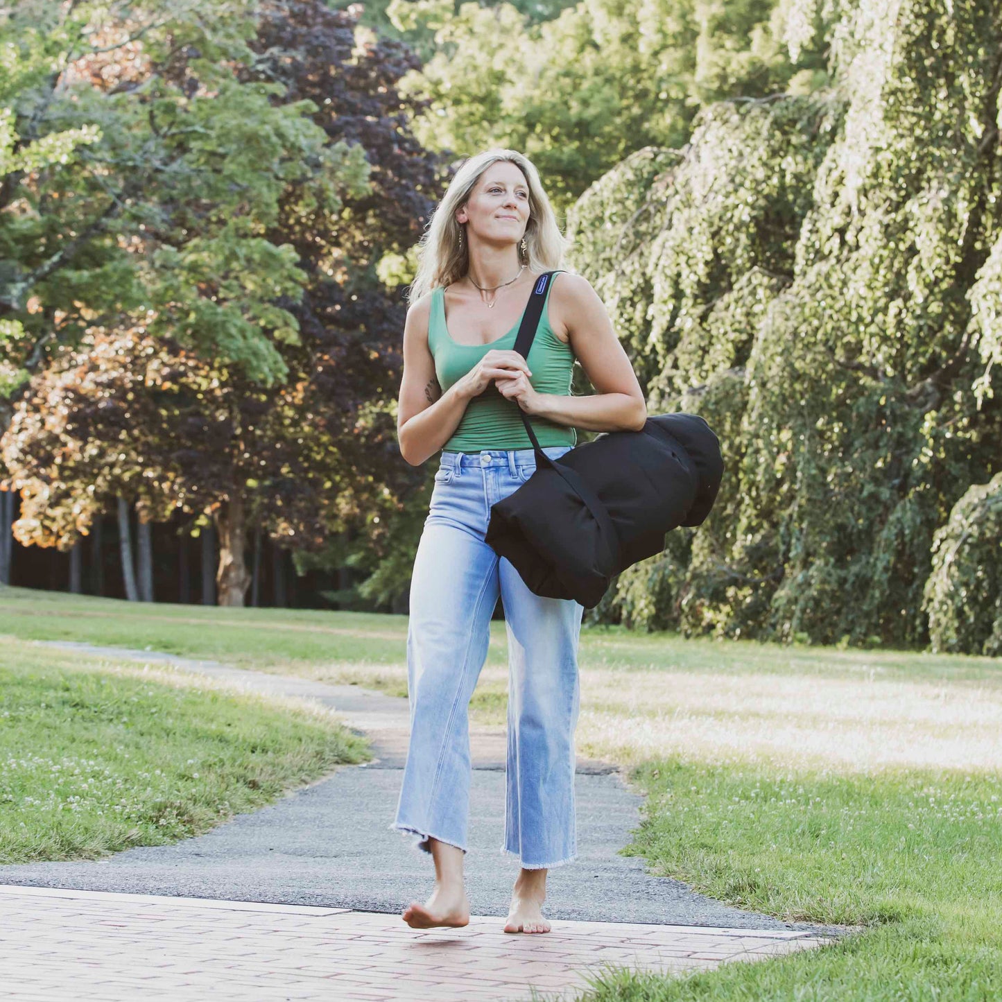 A woman walking barefoot on a paved path in a park, carrying a large black traveling meditation cushion over her shoulder.
