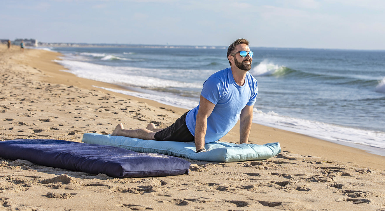 A man in a blue t-shirt and sunglasses does a yoga pose on a light blue cushion on a sandy beach, with a dark blue cushion nearby and the ocean in the background.