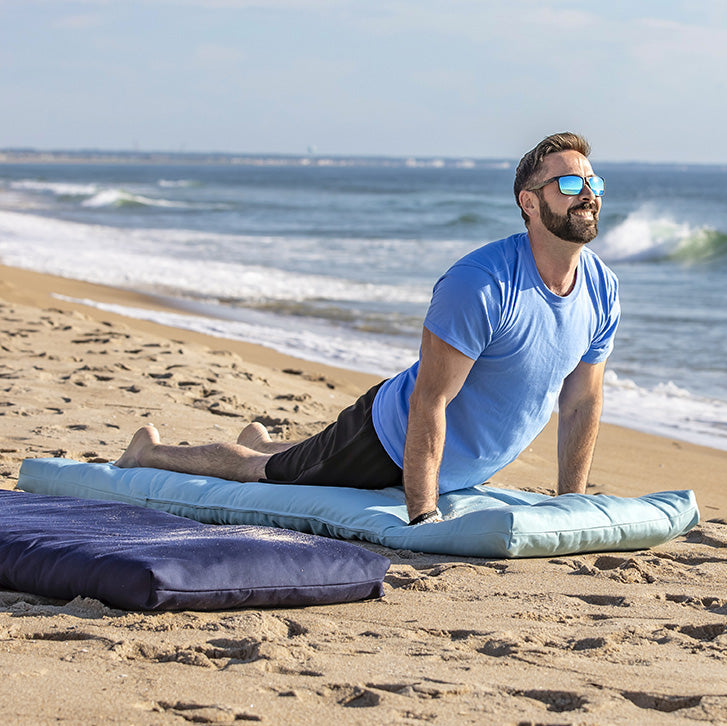 A man in a blue t-shirt and sunglasses does a yoga pose on a light blue cushion on a sandy beach, with a dark blue cushion nearby and the ocean in the background.