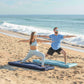 A man and a woman practice yoga on beach mats on sandy shore with ocean waves in the background.
