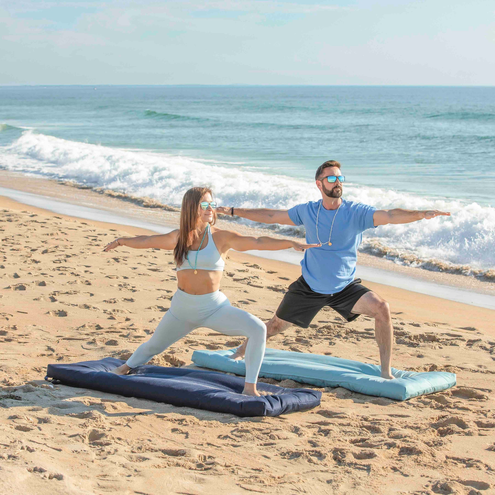 A man and a woman practice yoga on beach mats on sandy shore with ocean waves in the background.
