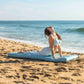 A woman in a blue yoga outfit is doing a yoga pose on a light blue beach mat on a sandy beach with the ocean in the background.