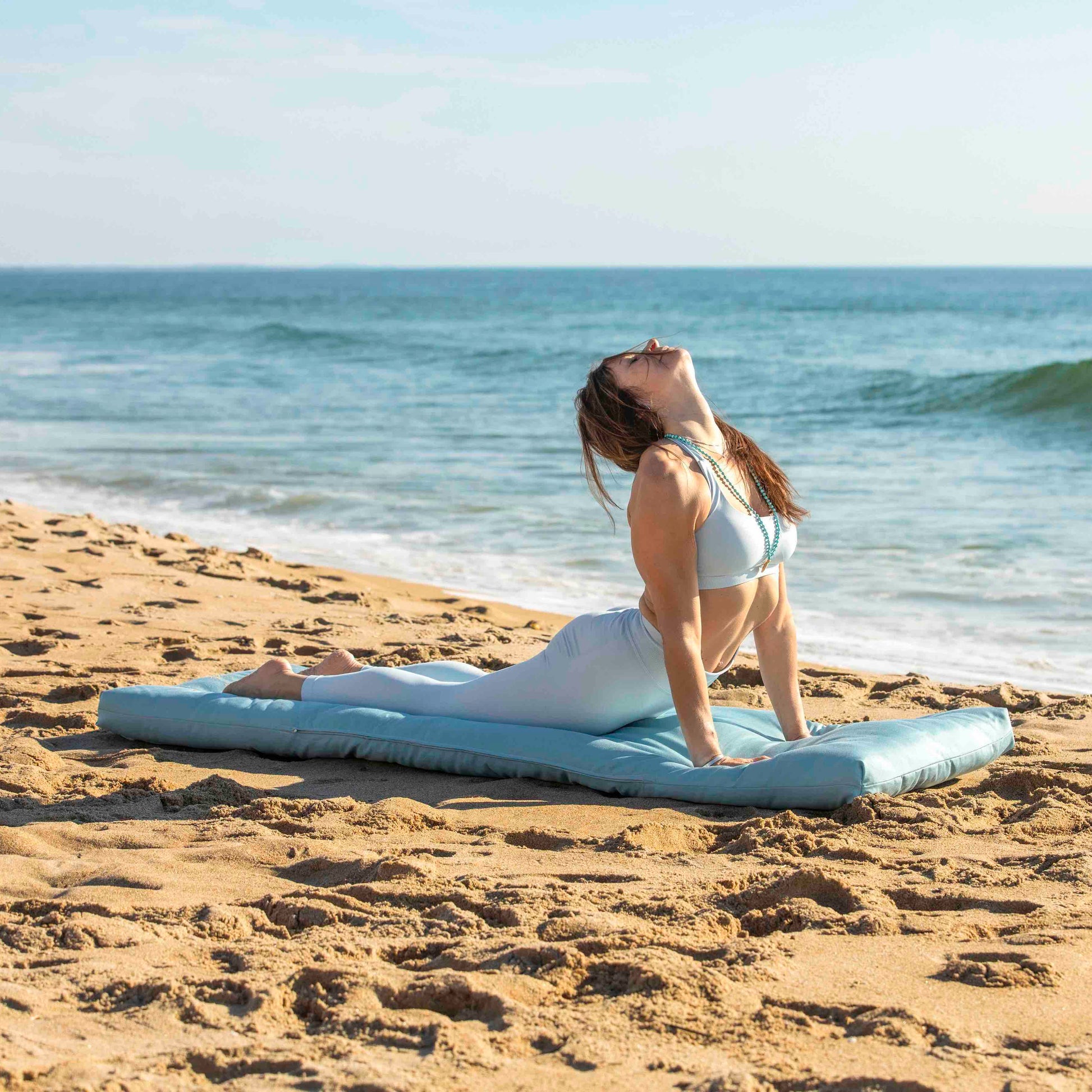 A woman in a blue yoga outfit is doing a yoga pose on a light blue beach mat on a sandy beach with the ocean in the background.