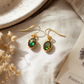 A pair of gold drop earrings with oval green iridescent stones, displayed on a textured cream linen fabric with dried flowers and a ceramic plate.