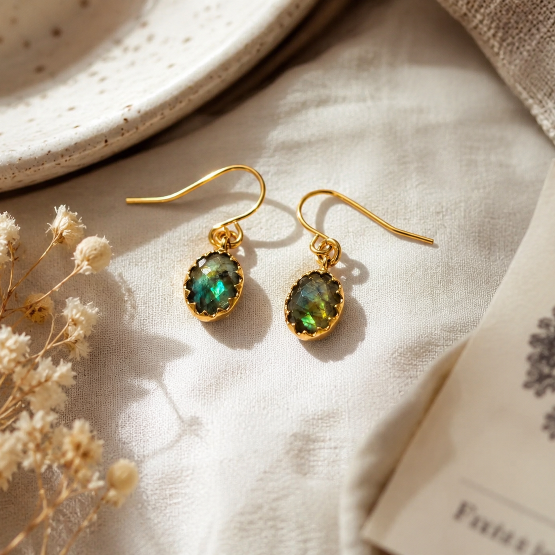 A pair of gold drop earrings with oval green iridescent stones, displayed on a textured cream linen fabric with dried flowers and a ceramic plate.