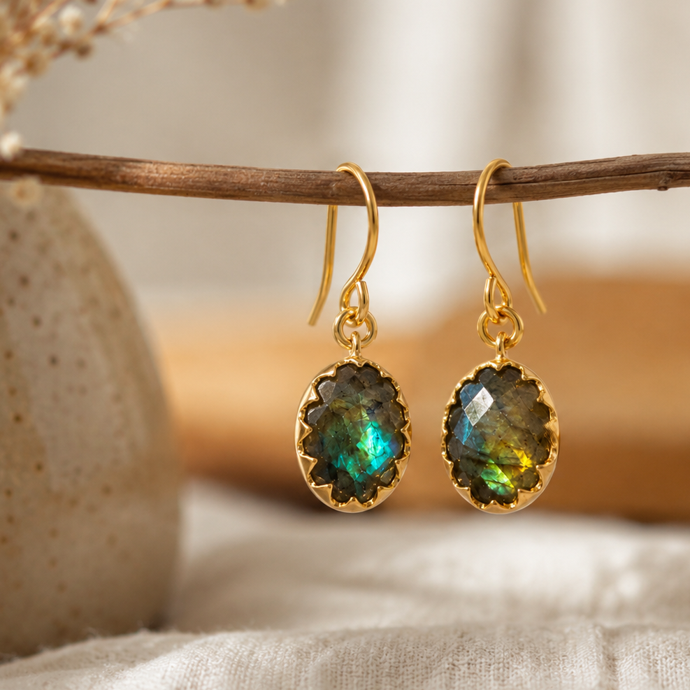 A pair of gold drop earrings with oval iridescent green-blue stones, hanging from a thin wooden branch in a styled setting with soft neutral fabric and dried floral decor.