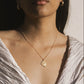 A close-up lifestyle shot of a model wearing a delicate gold chain necklace with a circular compass pendant, styled with a white draped top.