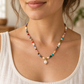 A close-up of a woman wearing a delicate beaded necklace with a large white pearl pendant. The necklace features small multicolored beads interspersed with white pearls and gold-toned accents, worn over a white ribbed tank top.