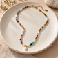 A beaded necklace with colorful stone-like beads, white pearl-like beads, and gold-toned accents, displayed on a round beige ceramic plate.